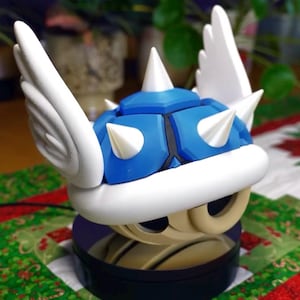 May include: A blue and white sculpture of a video game item, featuring a spiky shell with white wings. The sculpture is on a black base, set against a festive background with a Christmas theme.