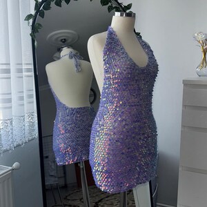 May include: A lavender sequin halter dress on a mannequin. The dress has a deep V-neck and a low back. The short dress ends above the knees. A mirror reflects the dress and a second mannequin. The background includes a white dresser and a decorative mirror.