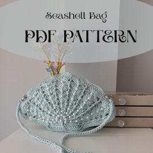 May include: A light blue crocheted seashell bag with pearl detailing and a matching strap. The bag is displayed on a white surface. The text "Seashell Bag PDF PATTERN" is at the top, with "english version" at the bottom.