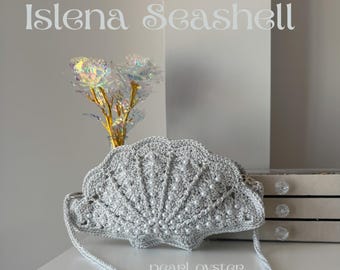May include: A silver crocheted seashell-shaped handbag with a braided strap. The bag is adorned with small white pearls and features the text "pearl oyster". The words "Islena Seashell" are above the bag.