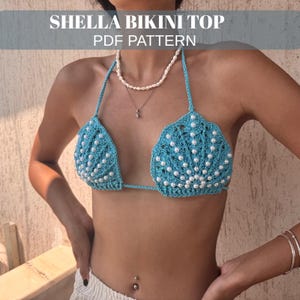 May include: A crocheted blue bikini top with white pearl embellishments. The top is in a shell design and has the text "SHELLA BIKINI TOP PDF PATTERN" at the top. The bikini top is a halter style.