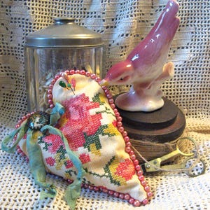 May include: A heart-shaped pincushion with a floral design and a blue ribbon. The pincushion is surrounded by pink beads and is sitting on a crocheted surface. There is a ceramic bird figurine and a spool of twine in the background.