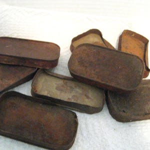 May include: A collection of seven rectangular metal tins with rounded corners, each showing significant oxidation and rust. The containers are shallow with low walls, displaying a weathered, dark brown, and metallic texture across their surfaces.