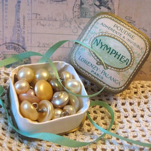 May include: A small square metal tin labeled POUDRE NYMPHEA LORENZY-PALANCA PARIS with its lid removed, filled with a collection of round, cream-colored buttons. A thin, light green ribbon is draped over the open tin and the buttons inside.