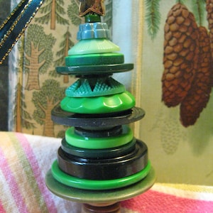 May include: A handmade Christmas tree ornament crafted from various green buttons, topped with a gold star. The tree is suspended by a teal ribbon with gold trim. The ornament is a unique, festive decoration.