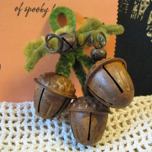 May include: A rustic Halloween decoration featuring three acorn-shaped bells with a rusty brown finish. The bells are attached to a green fuzzy stem. A bat silhouette and the words "of spooky" are visible in the background.