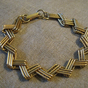 May include: A gold-colored bracelet with a woven design. The bracelet is made of multiple parallel bars that are interwoven to create a zig-zag pattern. The clasp is a simple hook and eye closure.