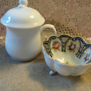 May include: A white porcelain sugar bowl with a lid and a small handle, next to a small, scalloped dish. The dish features a black and gold trim with painted roses. Both pieces are on a neutral-toned surface.