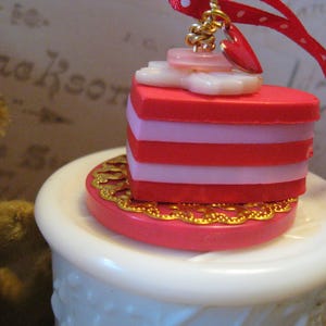 May include: A heart-shaped ornament with red and pink layers, topped with a small heart and button accents. The ornament is suspended by a red ribbon with white polka dots. The base has a gold filigree design.