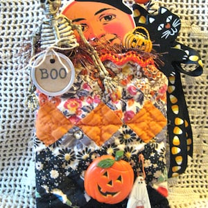 May include: A quilted fabric pouch featuring a diamond pattern in orange, black, and floral prints. It is decorated with a skeleton, a vintage-style moon face in a top hat, a black cat, a plastic pumpkin, a ghost figure, and a tag reading BOO.