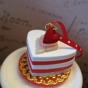 May include: A heart-shaped decorative item with a red and white layered design, topped with a red heart charm and ribbon. The item sits on a red plate with gold filigree edging. A charming, romantic accent.