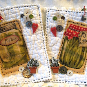 May include: Two quilted fabric squares with jar illustrations. One says "Pickles," the other "Dilly Beans." Each has decorative buttons, a heart, and a small pot with "Herbs." A mushroom and heart charm hang from the top.