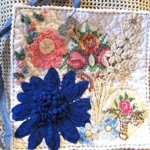 May include: A square white quilted fabric piece decorated with a large blue flower, a pink crocheted flower with a button center, a silver heart charm, cream lace floral appliqués, and two small cross-stitched floral motifs with blue ribbon ties.