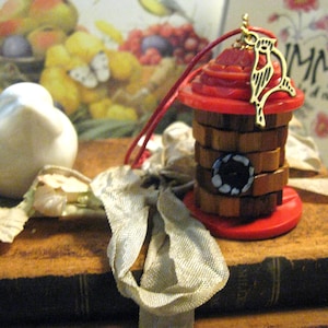 May include: A decorative birdhouse ornament with a red top and base, and a wooden body. The birdhouse has a gold-colored bird charm and a black button with white accents. A red string is attached for hanging.