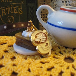 May include: A miniature charm featuring a stack of pancakes with whipped cream, a gold-colored charm with a leaf design, and a small white teapot with blue and red stripes. The items rest on a yellow crocheted surface.