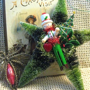 May include: A vintage Christmas ornament featuring a green, white, and red wooden figure wearing a hat and holding a red heart. The figure is surrounded by green and gold tinsel, a small green Christmas tree, and red berries.