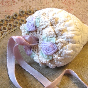 May include: A cream-colored, seashell-shaped decorative item with a crocheted top, adorned with lavender fabric roses, light green accents, and faux pearls. A pink ribbon is attached for hanging.