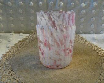 May include: A clear glass vase with a pink and white abstract design. The vase is cylindrical with a slightly flared top. It sits on a tan placemat with a decorative lace edge. The background is a white fabric with a dotted pattern.