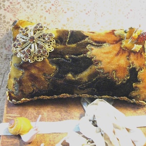 May include: A rectangular velvet pincushion in shades of brown and gold, featuring a decorative gold-tone brooch with amber-colored stones on the left side and a cluster of pins on the right, finished with a gold braided trim along the edges.
