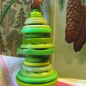 May include: A handmade Christmas tree ornament crafted from graduated green buttons, topped with a gold star. The tree is a vibrant mix of green shades, creating a festive and unique holiday decoration. The ornament is approximately 4 inches tall.