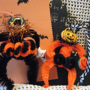 May include: Two Halloween-themed pipe cleaner figures. One is a black cat spider with orange accents and the other is an orange pumpkin figure with a jack-o'-lantern face. Both have decorative tinsel and are on a Halloween-themed background.