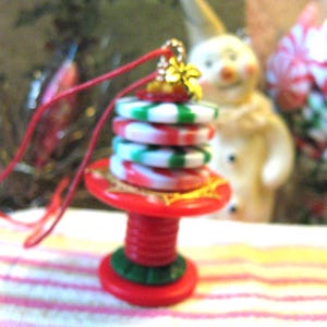 May include: A Christmas ornament shaped like a spool of thread. The ornament is red, green, and white, with a gold-colored top and a red string for hanging. The ornament has a festive, holiday theme.