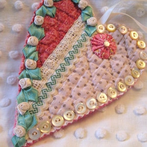 May include: Handmade quilted heart ornament with a ribbon for hanging. The heart features a patchwork design with red and pink fabrics, lace, and decorative buttons. Accented with green ribbon and small fabric roses.