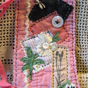 May include: A rectangular pink fabric bookmark featuring layered patches of black, red, and white fabric. It is decorated with a white dragonfly charm, embroidered green leaves, a white flower, a pansy, and four clear buttons with black stitching.