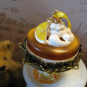 May include: A decorative bottle stopper with a lemon slice charm. The stopper features a brown base, white frosting-like details, and a yellow ribbon. The charm is a yellow lemon slice with white accents.