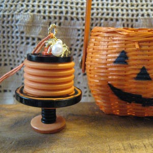 May include: A miniature Halloween-themed display featuring a stack of orange buttons on a black and orange stand, topped with a spider and pearl embellishment. A woven orange pumpkin basket with a jack-o'-lantern face is in the background.