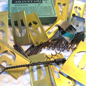 May include: A collection of vintage brass stencils with cut-out letters and numbers, scattered around a green box labeled "ADJUSTABLE STENCILS". Includes a heart-shaped cookie cutter and a metal tool with a twisted handle. The stencils are for lettering jobs.