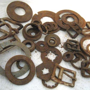 May include: A collection of assorted metal hardware components including various circular washers, flat rings, and rectangular belt buckles. These items exhibit a weathered, oxidized brown rust patina across their surfaces, showing signs of age and wear.