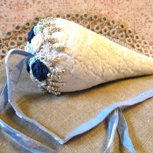 May include: A white, cone-shaped item with a textured surface, adorned with blue and white fabric flowers, silver beads, and a light blue ribbon. The item is resting on a tan fabric surface, suggesting a handcrafted or decorative item.