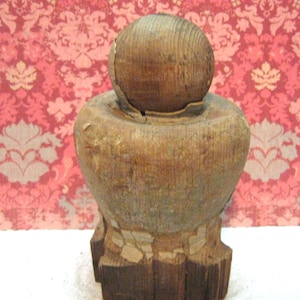 May include: A weathered wooden finial featuring a spherical top resting on a rounded, tapered body. The base is square-shaped with visible chipped paint and exposed wood grain, showing signs of age and wear throughout the entire structure.