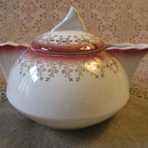 May include: A white ceramic teapot with a lid, featuring a red and gold floral design. The teapot has a unique shape with a pointed spout and a decorative handle. The rim and lid are accented with a deep red color.