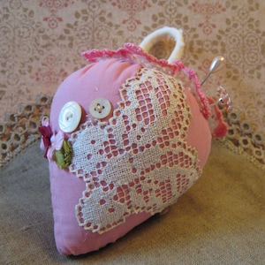 May include: A pink heart-shaped pincushion with white lace detailing and two white buttons. The pincushion has a small loop at the top and is decorated with small pink flowers and a pin. The background is a floral pattern.