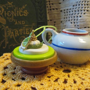 May include: A decorative bottle stopper with a lime slice charm, stacked on green, white, and brown layers. A small white teapot with blue and red accents sits nearby. The background includes a book titled "Picnics and Parties."