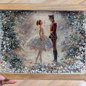 May include: A framed artwork featuring a ballet scene with a ballerina and a soldier in a winter setting. The artwork is encased in resin with glitter and small stones, and framed in a light-colored wood.