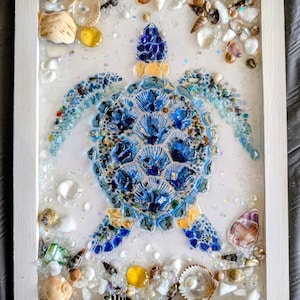 May include: A rectangular white-framed wall art piece featuring a sea turtle mosaic crafted from blue glass shards, small shells, and pebbles. The turtle is surrounded by scattered white shells, clear glass beads, and amber stones on a white background.