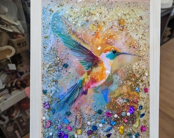 May include: A framed artwork featuring a vibrant hummingbird in flight. The bird has colorful plumage with shades of orange, blue, and green. The artwork is embellished with glitter and colorful gems, set within a white wooden frame.