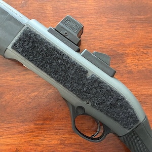 May include: Close-up of a gray and black firearm with a red dot sight. The firearm has a textured grip and a black, rectangular attachment. The red dot sight is mounted on top and has two adjustment dials.
