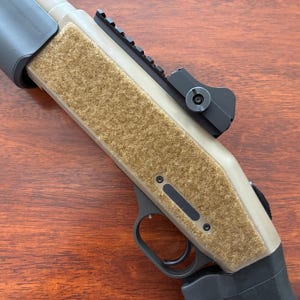 May include: A tan and black firearm with a textured, tan grip area. The firearm has a black rail system and a black sight. The trigger and other components are black. The firearm is angled, showing its side.