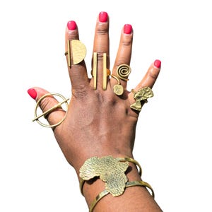 May include: A hand displaying various gold-toned rings and a bracelet. The rings feature geometric shapes, spirals, and a bust. The bracelet is shaped like the African continent. The nails are painted a bright pink.