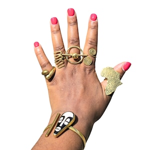 May include: A hand displaying various gold-toned rings and a bracelet. The rings feature unique designs, including spirals, a circle with a sphere, and an African continent shape. The bracelet has a black and white mask design.