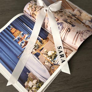 May include: A white satin ribbon tied in a bow, with the name "SARA" printed on one tail, rests on an open magazine. The magazine features images of a tiered cake, floral arrangements, and table settings, suggesting a wedding or event theme.