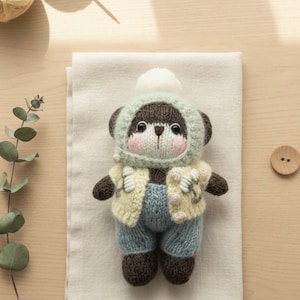 May include: A handmade knitted bear doll wearing a light blue hooded hat, a yellow vest, and blue pants. The bear has brown arms and legs, and a white pom-pom on the hat. The doll is sitting on a white cloth.