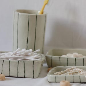 Puede incluir: Un conjunto de accesorios de baño de cerámica, que incluye un porta cepillos de dientes con un cepillo de dientes amarillo, dos bandejas rectangulares y un cuenco pequeño. Los artículos son de color blanco roto con rayas verticales verde oscuro.