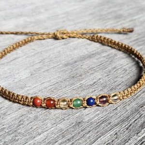 May include: A brown braided cord bracelet with seven small beads in the colours of the rainbow. The beads are red, orange, yellow, green, blue, indigo, and violet.