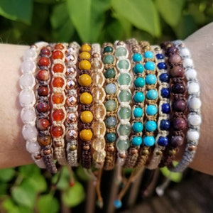 May include: A set of 12 colourful beaded bracelets with brown cord. The bracelets feature various gemstones, including white, red, yellow, green, blue, and purple.