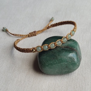 May include: A brown string bracelet with light green beads and gold accents. The bracelet is tied around a green stone.